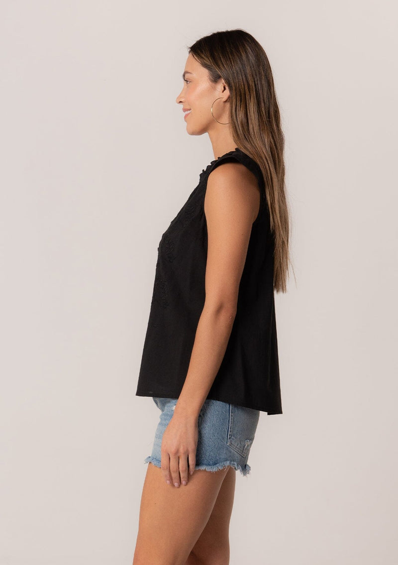[Color: Black] A side facing image of a brunette model wearing a bohemian cotton spring top in a black colorway. With short cap sleeves, a self covered button front, a ruffled neckline, a v neckline, and embroidered detail.