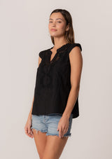[Color: Black] An angled front facing image of a brunette model wearing a bohemian cotton spring top in a black colorway. With short cap sleeves, a self covered button front, a ruffled neckline, a v neckline, and embroidered detail.