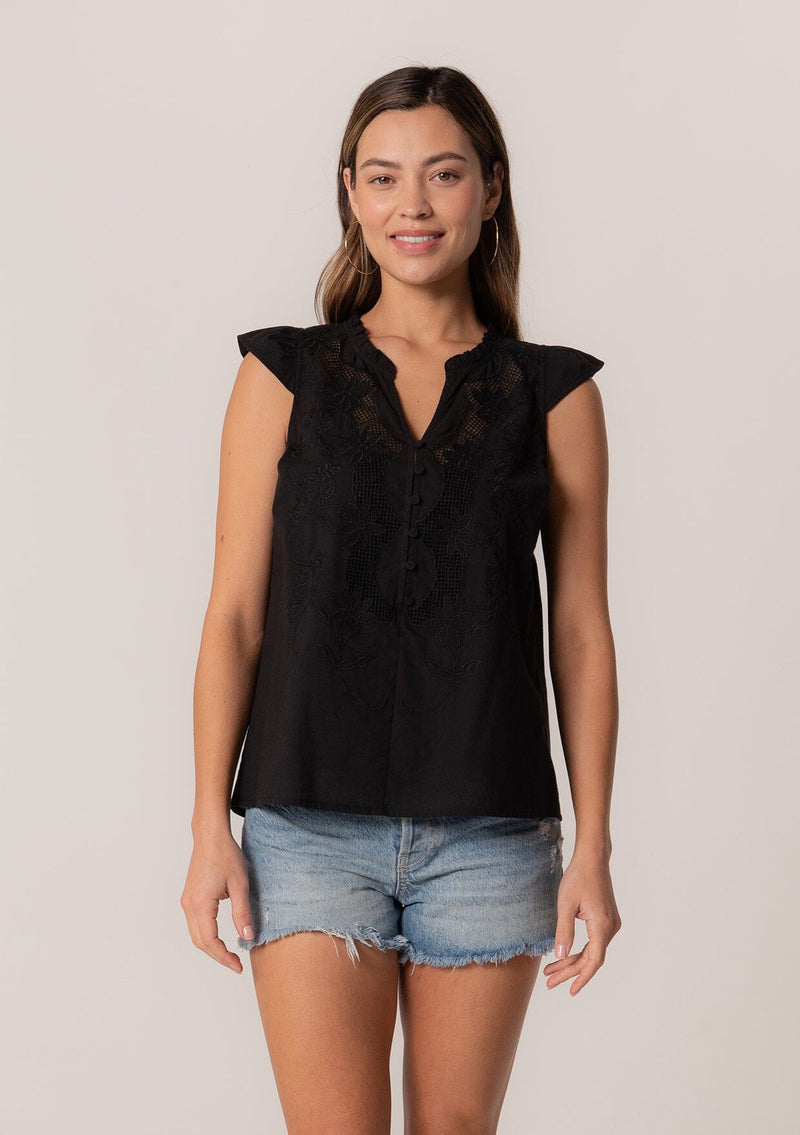 [Color: Black] A front facing image of a brunette model wearing a bohemian cotton spring top in a black colorway. With short cap sleeves, a self covered button front, a ruffled neckline, a v neckline, and embroidered detail.