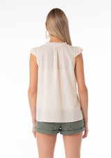 [Color: Natural] A back facing image of a blonde model wearing a bohemian cotton spring top in a natural colorway. With short cap sleeves, a self covered button front, a ruffled neckline, a v neckline, and embroidered detail. 