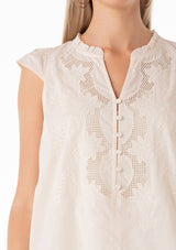[Color: Natural] A close up front facing image of a blonde model wearing a bohemian cotton spring top in a natural colorway. With short cap sleeves, a self covered button front, a ruffled neckline, a v neckline, and embroidered detail. 
