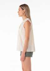 [Color: Natural] A side facing image of a blonde model wearing a bohemian cotton spring top in a natural colorway. With short cap sleeves, a self covered button front, a ruffled neckline, a v neckline, and embroidered detail. 