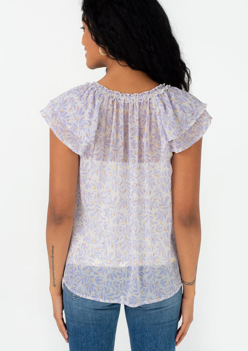 [Color: Natural/Lilac] A back facing image of a brunette model wearing a pretty spring blouse in a metallic clip dot chiffon purple leaf print design. With short flutter sleeves, a ruffled neckline, a split v neckline with ties, and a relaxed fit. 