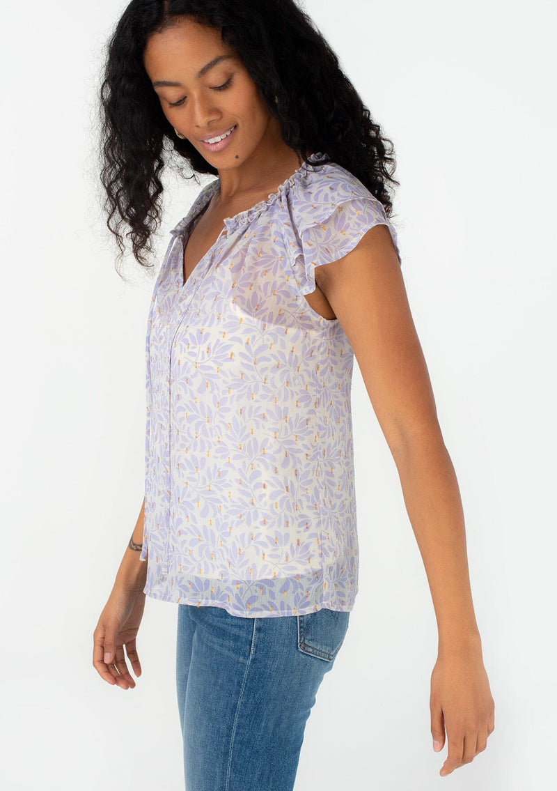 [Color: Natural/Lilac] A side facing image of a brunette model wearing a pretty spring blouse in a metallic clip dot chiffon purple leaf print design. With short flutter sleeves, a ruffled neckline, a split v neckline with ties, and a relaxed fit. 
