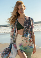 [Color: Natural/Coral] A half body front facing image of a blonde model on the beach wearing a lightweight bohemian kimono in a natural and coral mixed floral print. With half length kimono sleeves, an open front, and side slits.