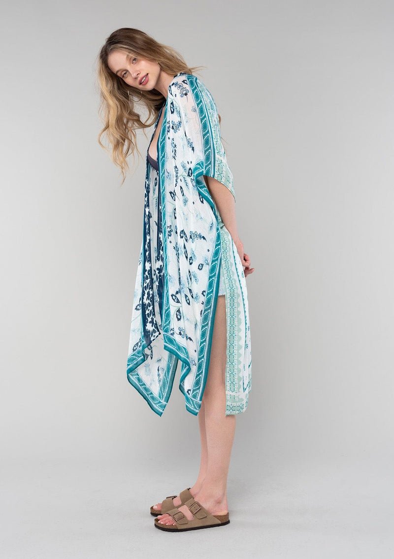 [Color: Ivory/Teal] A side facing image of a blonde model wearing a mid length bohemian kimono in an ivory and teal paisley and floral print. With half length kimono sleeves, an open front, and side slits. 
