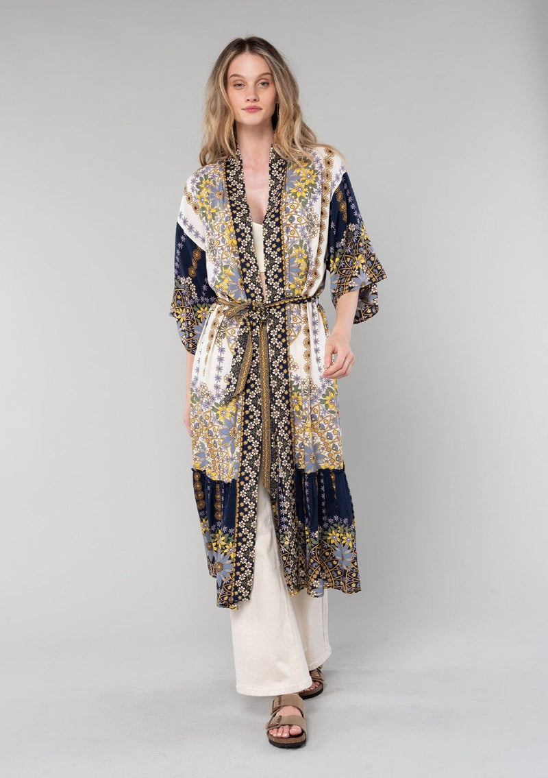 [Color: Ivory/Lilac] A front facing image of a blonde model wearing a bohemian duster lounge robe in an ivory, blue, and lilac purple patchwork floral print. With half length sleeves, a ruffle trimmed tiered hemline, and a tie waist belt. 