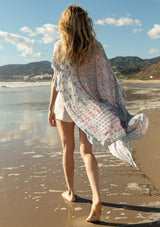 [Color: Ivory/Light Rose] A back facing image of a blonde model on the beach wearing a lightweight bohemian mid length kimono in an ivory and light pink mixed floral border print. With half length kimono sleeves, an open front, and side slits.