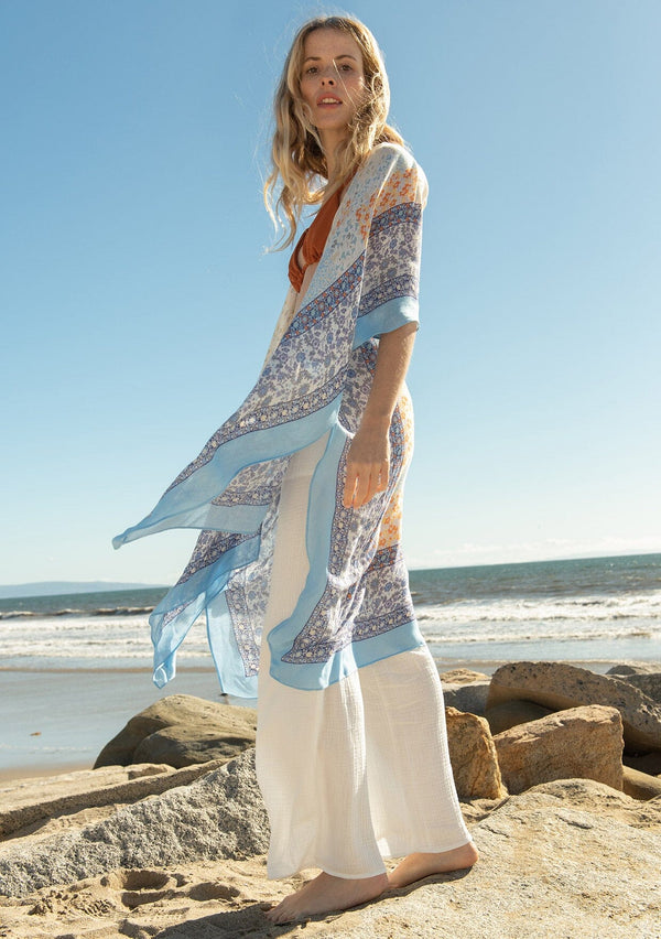 [Color: Ivory/Light Blue] A full body side facing image of a blonde model on the beach wearing a lightweight bohemian mid length kimono in an ivory and light blue mixed floral border print. With half length kimono sleeves, an open front, and side slits.