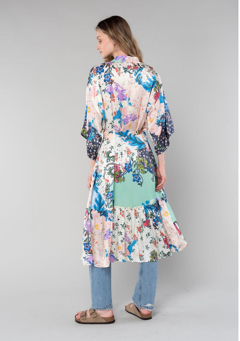 [Color: Ivory/Grey] A back facing image of a blonde model wearing a soft and silky bohemian lounge duster kimono robe in a multi color floral patchwork print. With half length kimono sleeves, a ruffle trimmed tiered hemline, and a tie waist belt. 