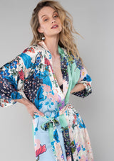 [Color: Ivory/Grey] A close up front facing image of a blonde model wearing a soft and silky bohemian lounge duster kimono robe in a multi color floral patchwork print. With half length kimono sleeves, a ruffle trimmed tiered hemline, and a tie waist belt. 