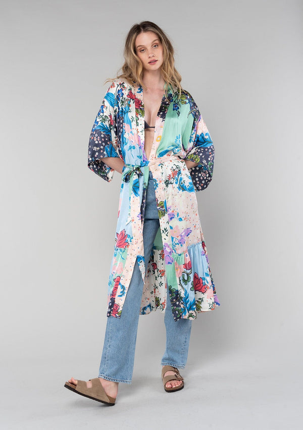 [Color: Ivory/Grey] A front facing image of a blonde model wearing a soft and silky bohemian lounge duster kimono robe in a multi color floral patchwork print. With half length kimono sleeves, a ruffle trimmed tiered hemline, and a tie waist belt. 