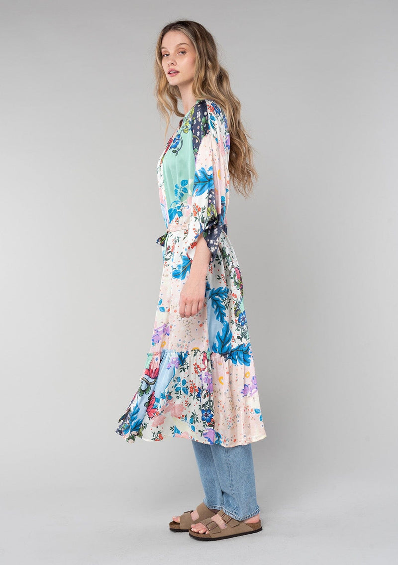 [Color: Ivory/Grey] A side facing image of a blonde model wearing a soft and silky bohemian lounge duster kimono robe in a multi color floral patchwork print. With half length kimono sleeves, a ruffle trimmed tiered hemline, and a tie waist belt. 