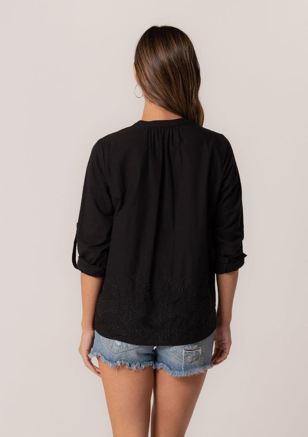 [Color: Black] A back facing image of a brunette model wearing a black cotton button front blouse with long rolled sleeves, a v neckline, embroidered details, and pintuck details along the yoke. 