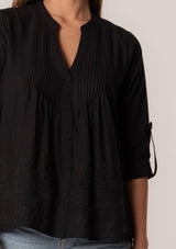 [Color: Black] A close up front facing image of a brunette model wearing a black cotton button front blouse with long rolled sleeves, a v neckline, embroidered details, and pintuck details along the yoke.
