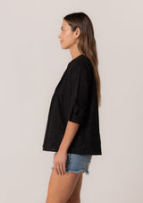 [Color: Black] A side facing image of a brunette model wearing a black cotton button front blouse with long rolled sleeves, a v neckline, embroidered details, and pintuck details along the yoke.