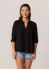[Color: Black] A front facing image of a brunette model wearing a black cotton button front blouse with long rolled sleeves, a v neckline, embroidered details, and pintuck details along the yoke.