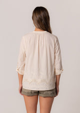 [Color: Natural] A back facing image of a brunette model wearing an ivory cotton button front blouse with long rolled sleeves, a v neckline, embroidered details, and pintuck details along the yoke.