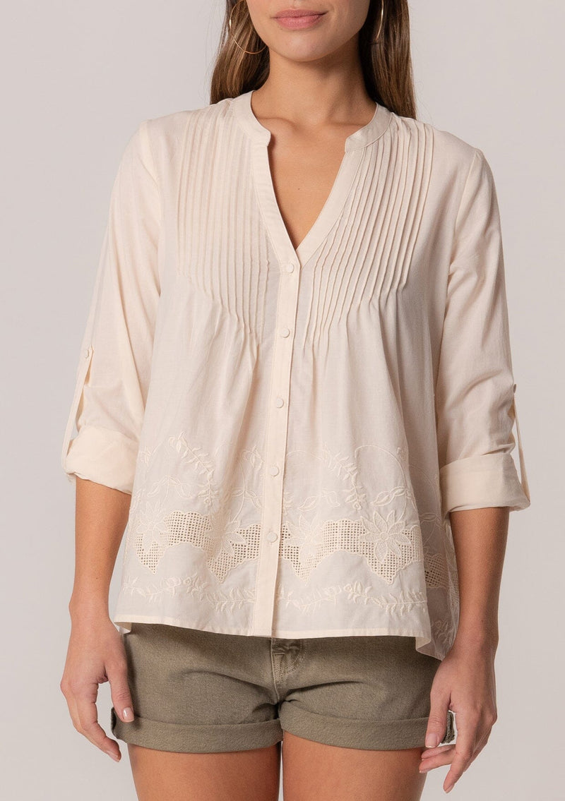 [Color: Natural] A close up front facing image of a brunette model wearing an ivory cotton button front blouse with long rolled sleeves, a v neckline, embroidered details, and pintuck details along the yoke.