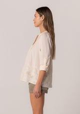 [Color: Natural] A side facing image of a brunette model wearing an ivory cotton button front blouse with long rolled sleeves, a v neckline, embroidered details, and pintuck details along the yoke.