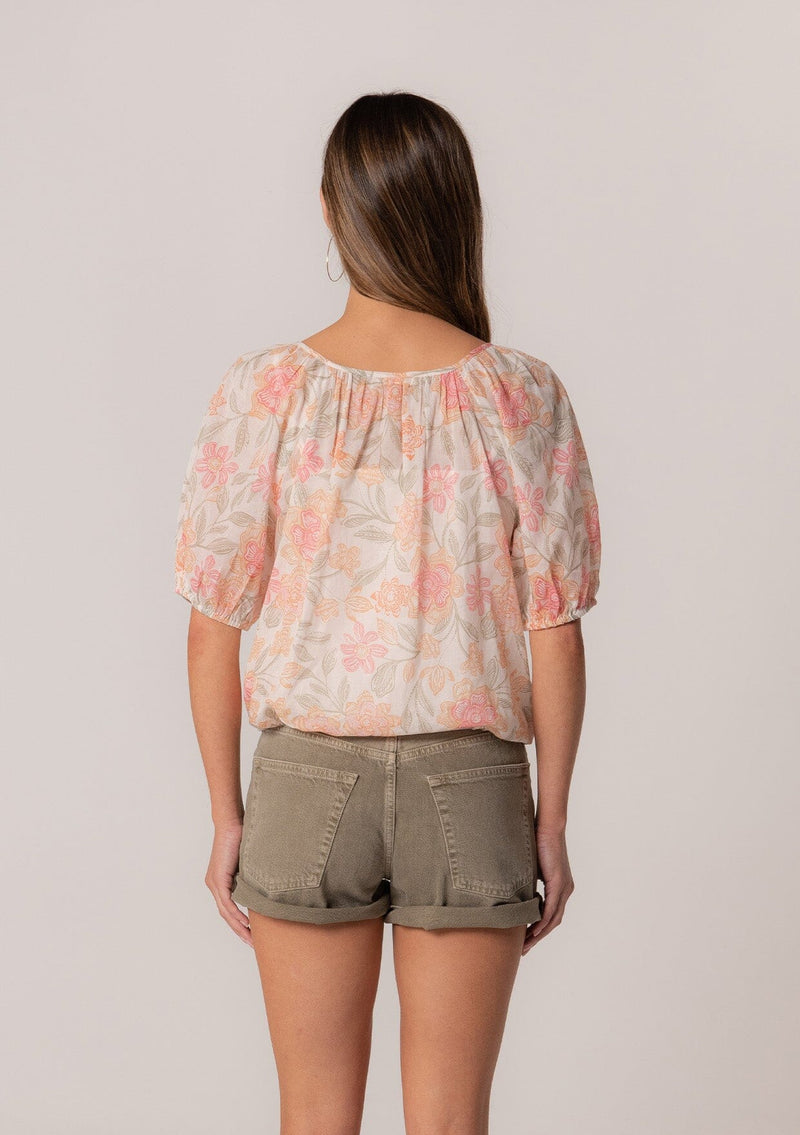 [Color: Natural/Coral] A back facing image of a brunette model wearing a cotton summer blouse in a pink floral print. With short puff sleeves, a button front, a scoop neckline, and a drawstring waist with front ties. 