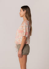 [Color: Natural/Coral] A side facing image of a brunette model wearing a cotton summer blouse in a pink floral print. With short puff sleeves, a button front, a scoop neckline, and a drawstring waist with front ties. 