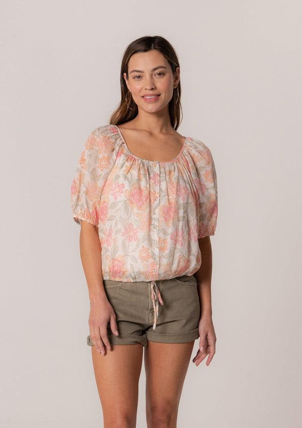 [Color: Natural/Coral] A front facing image of a brunette model wearing a cotton summer blouse in a pink floral print. With short puff sleeves, a button front, a scoop neckline, and a drawstring waist with front ties. 