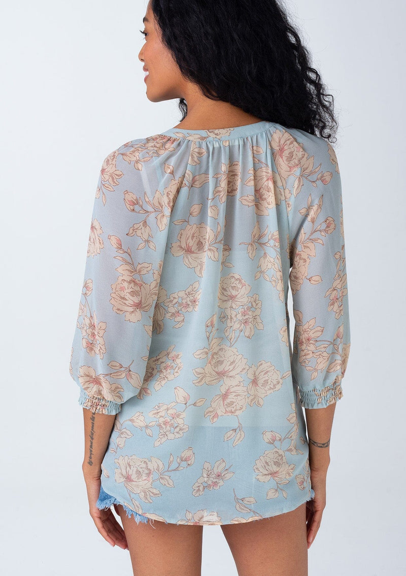 [Color: Dusty Blue/Natural] A back facing image of a brunette model wearing a bohemian spring sheer chiffon blouse in a dusty blue and natural floral print. With three quarter length sleeves, a button front, and a split v neckline. 