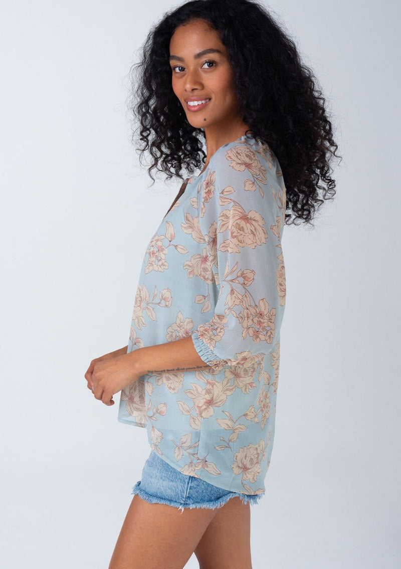 [Color: Dusty Blue/Natural] A side facing image of a brunette model wearing a bohemian spring sheer chiffon blouse in a dusty blue and natural floral print. With three quarter length sleeves, a button front, and a split v neckline. 