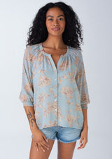 [Color: Dusty Blue/Natural] A front facing image of a brunette model wearing a bohemian spring sheer chiffon blouse in a dusty blue and natural floral print. With three quarter length sleeves, a button front, and a split v neckline. 
