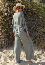 [Color: Olive] A woman standing outside on the beach wearing a cropped wide leg pant. Featuring an elastic waistband and an embroidered trim along the side.
