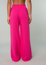 [Color: Fuchsia] A back facing image of a brunette model wearing a bright pink resort lounge pant in cotton gauze. With a wide leg, side pockets, and a smocked elastic waistband. 
