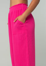 [Color: Fuchsia] A close up side facing image of a brunette model wearing a bright pink resort lounge pant in cotton gauze. With a wide leg, side pockets, and a smocked elastic waistband. 