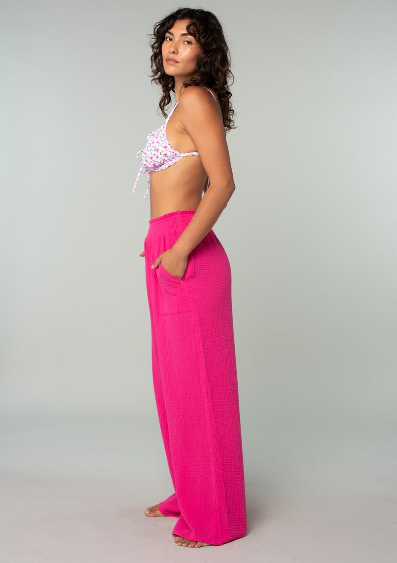 [Color: Fuchsia] A full body side facing image of a brunette model wearing a bright pink resort lounge pant in cotton gauze. With a wide leg, side pockets, and a smocked elastic waistband. 