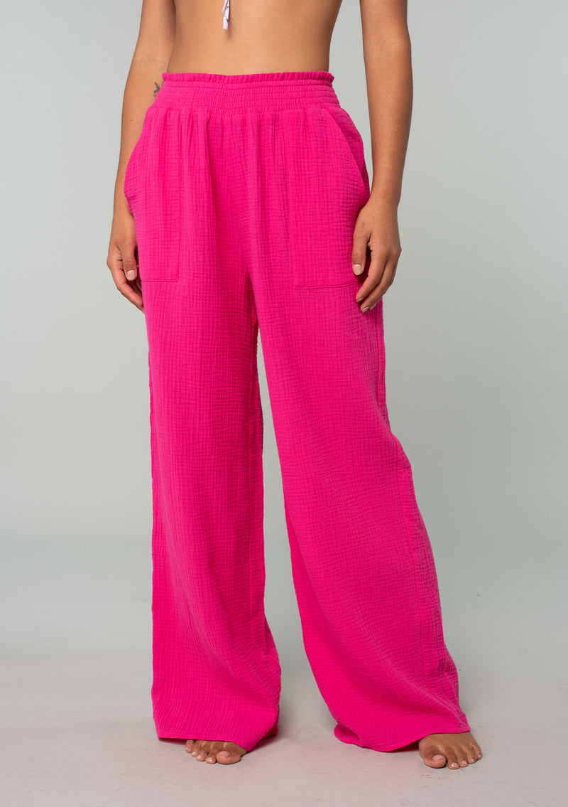 [Color: Fuchsia] A half body front facing image of a brunette model wearing a bright pink resort lounge pant in cotton gauze. With a wide leg, side pockets, and a smocked elastic waistband. 