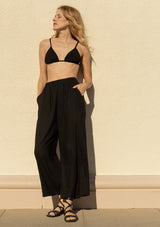 [Color: Black] A full body front facing image of a blonde model outside wearing a cool black linen and cotton blend lounge pant. With side pockets, an elastic waistband, and a wide leg.