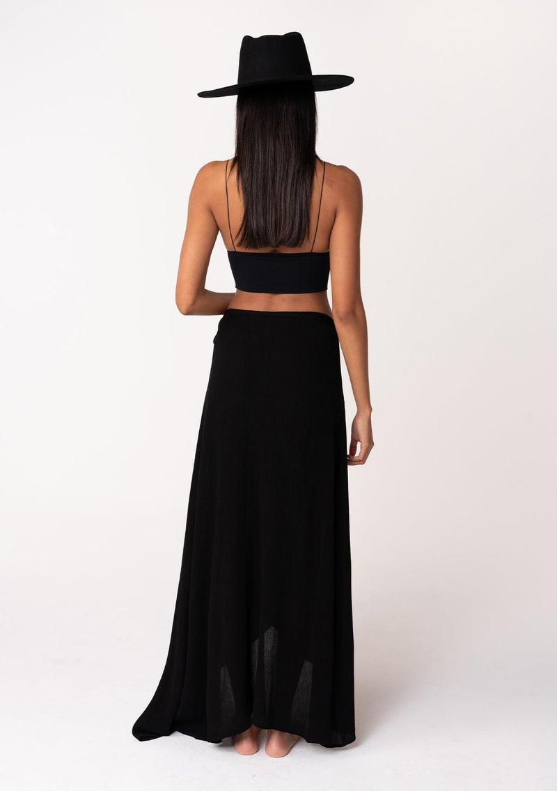 [Color: Black] A back facing image of a brunette model wearing a classic flowy black bohemian maxi wrap skirt with a slit and side tie closure. 