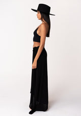 [Color: Black] A side facing image of a brunette model wearing a classic flowy black bohemian maxi wrap skirt with a slit and side tie closure. 
