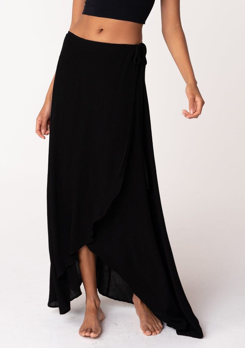 [Color: Black] A half body front facing image of a brunette model wearing a classic flowy black bohemian maxi wrap skirt with a slit and side tie closure. 