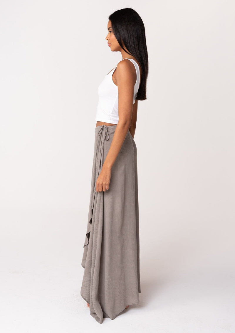 [Color: Cement] A side facing image of a brunette model wearing a classic flowy bohemian maxi wrap skirt with a slit and side tie closure. 