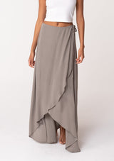 [Color: Cement] A half body front facing image of a brunette model wearing a classic flowy bohemian maxi wrap skirt with a slit and side tie closure. 