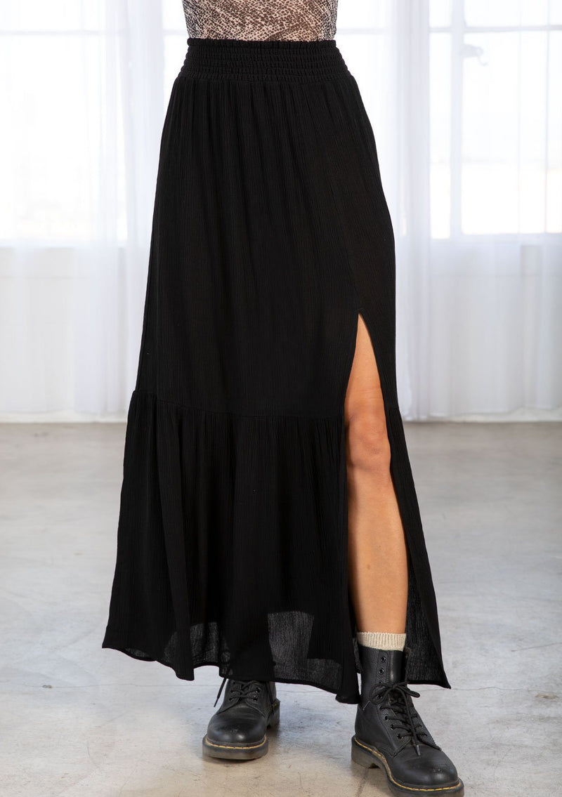 [Color: Black] A model wearing a black maxi skirt. With a smocked elastic waist, a side slit, a tiered skirt, and a flowy silhouette. 