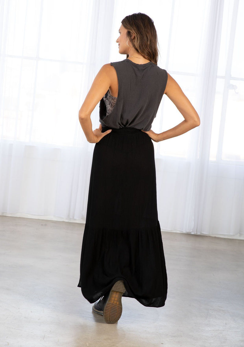 [Color: Black] A model wearing a black maxi skirt. With a smocked elastic waist, a side slit, a tiered skirt, and a flowy silhouette. 