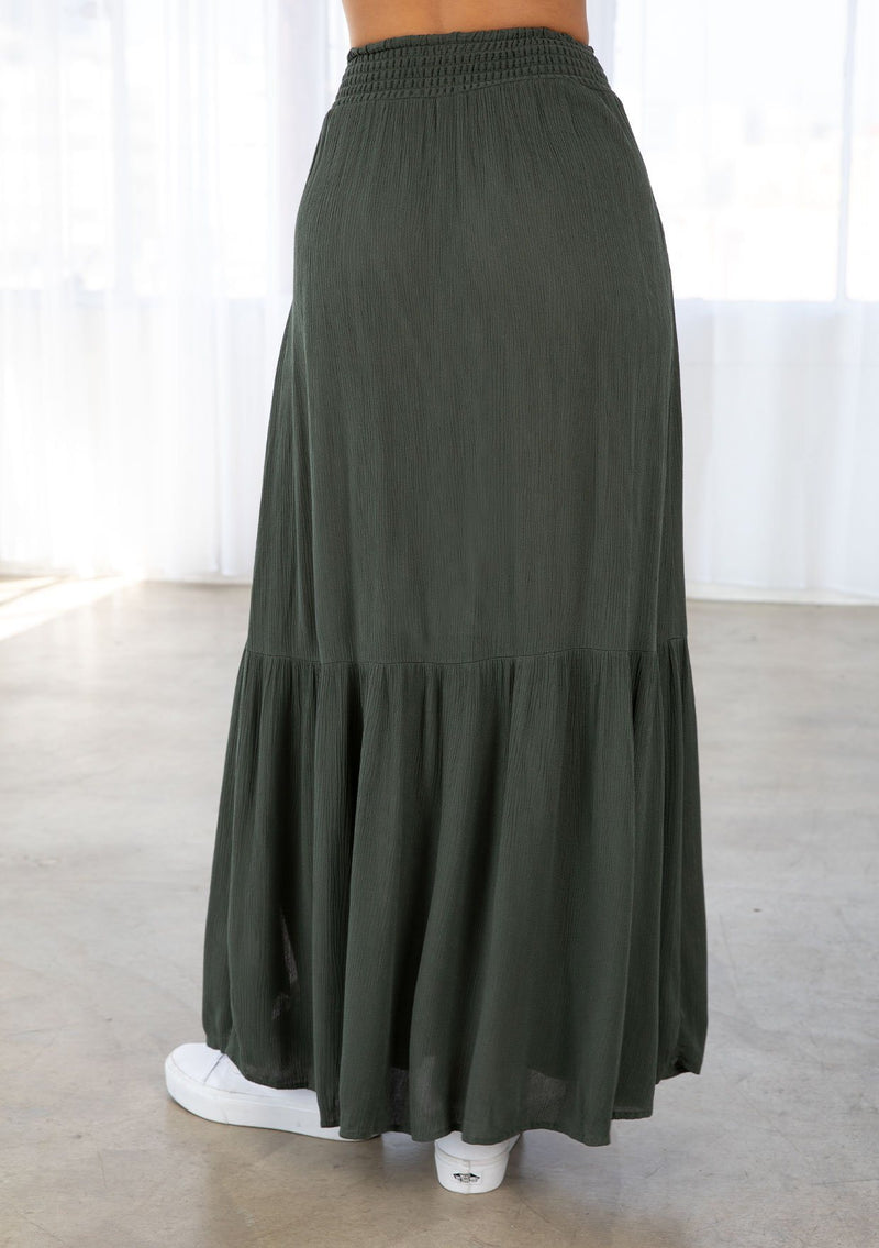 [Color: Military] A model wearing a military green maxi skirt. With a smocked elastic waist, a side slit, a tiered skirt, and a flowy silhouette. 