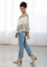 [Color: Oatmeal/Grey] A model wearing an oatmeal and grey poncho sweater in a novelty design. With contrast stitch details, pom pom neck ties, and a cropped length. 