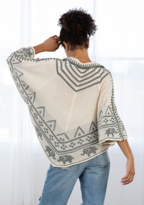 [Color: Oatmeal/Grey] A model wearing an oatmeal and grey poncho sweater in a novelty design. With contrast stitch details, pom pom neck ties, and a cropped length. 