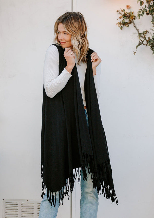 [Color: Black] A woman outside wearing a hooded knit fringe vest. Featuring a trending fringed hemline and an easy open front.