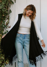 [Color: Black] A woman outside wearing a hooded knit fringe vest. Featuring a trending fringed hemline and an easy open front.