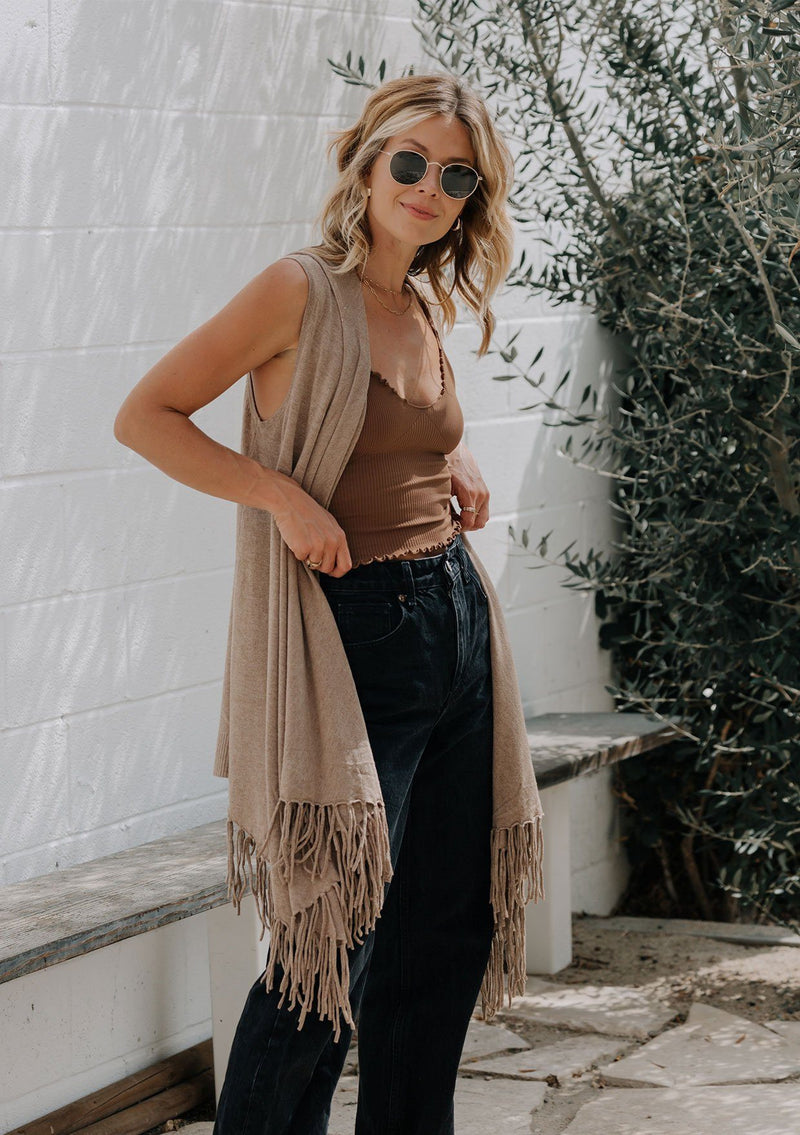 [Color: Heather Coco] A woman outside wearing a hooded knit fringe vest. Featuring a trending fringed hemline and an easy open front.