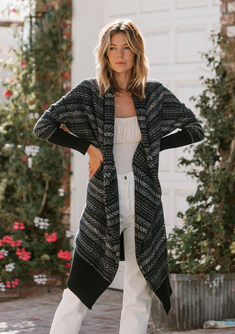 [Color: Black/Grey] A model wearing a cozy reversible shawl collar cardigan sweater in a black and grey southwestern inspired stripe jacquard. With long dolman sleeves and contrast ribbed trim. 